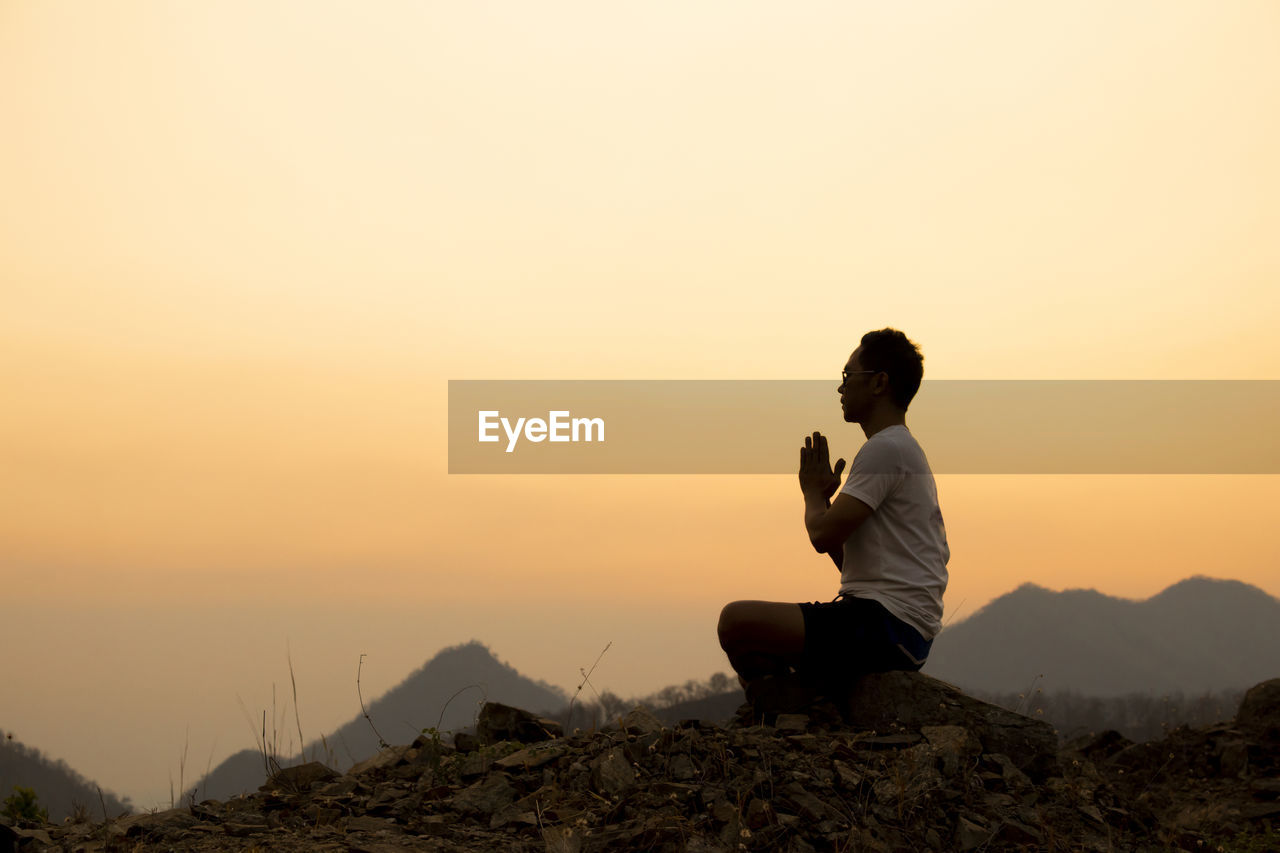 REAR VIEW OF MAN SITTING ON MOUNTAIN AGAINST SKY DURING SUNSET