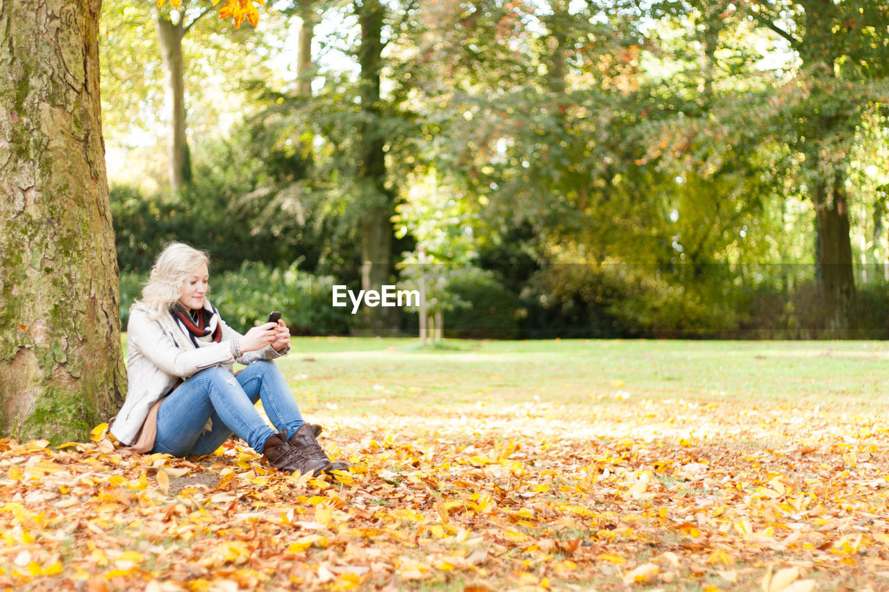 Woman using phone while sitting on fallen autumn leaves at park