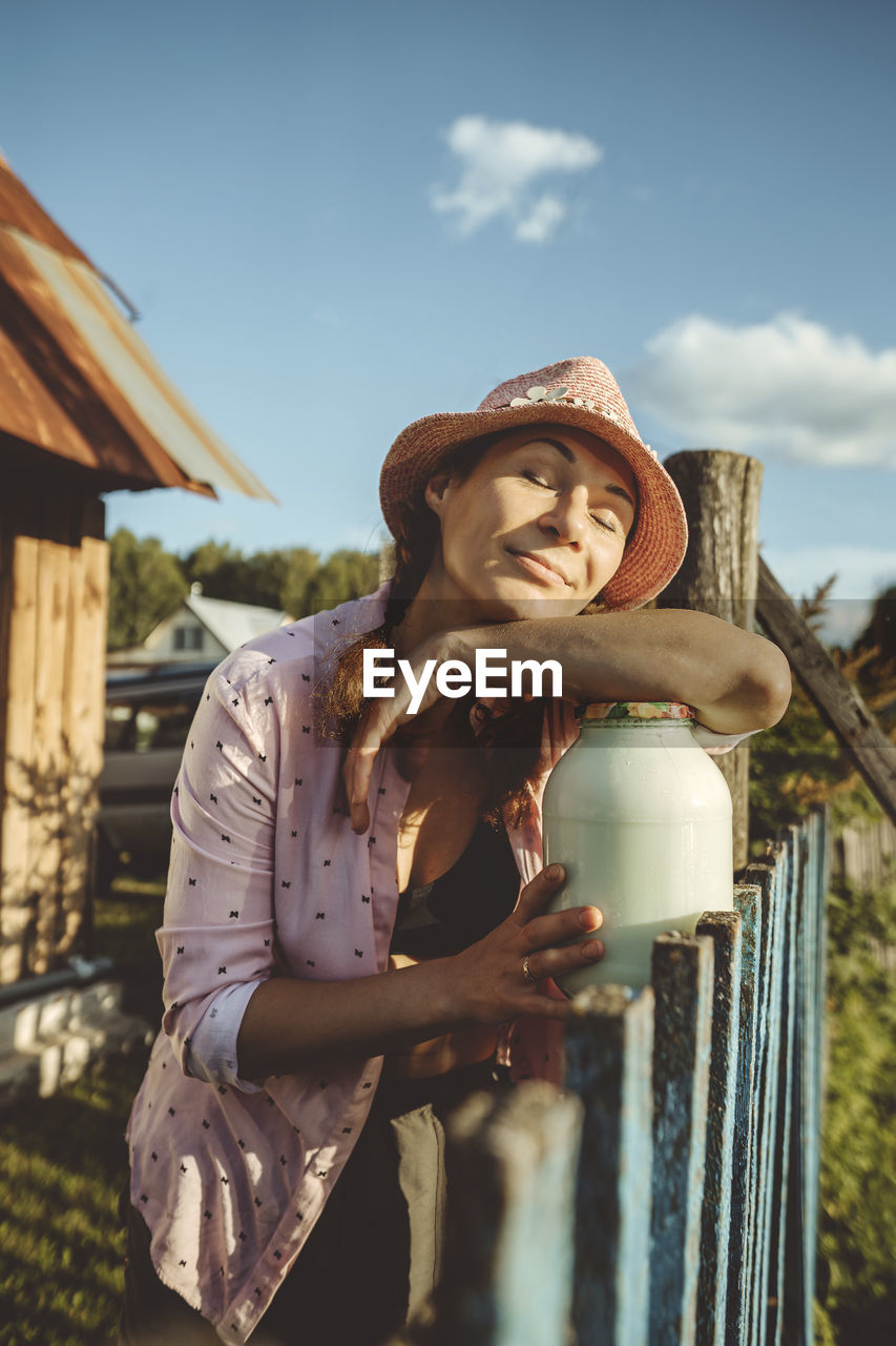 Woman with eyes closed leaning on jar full of milk by fence