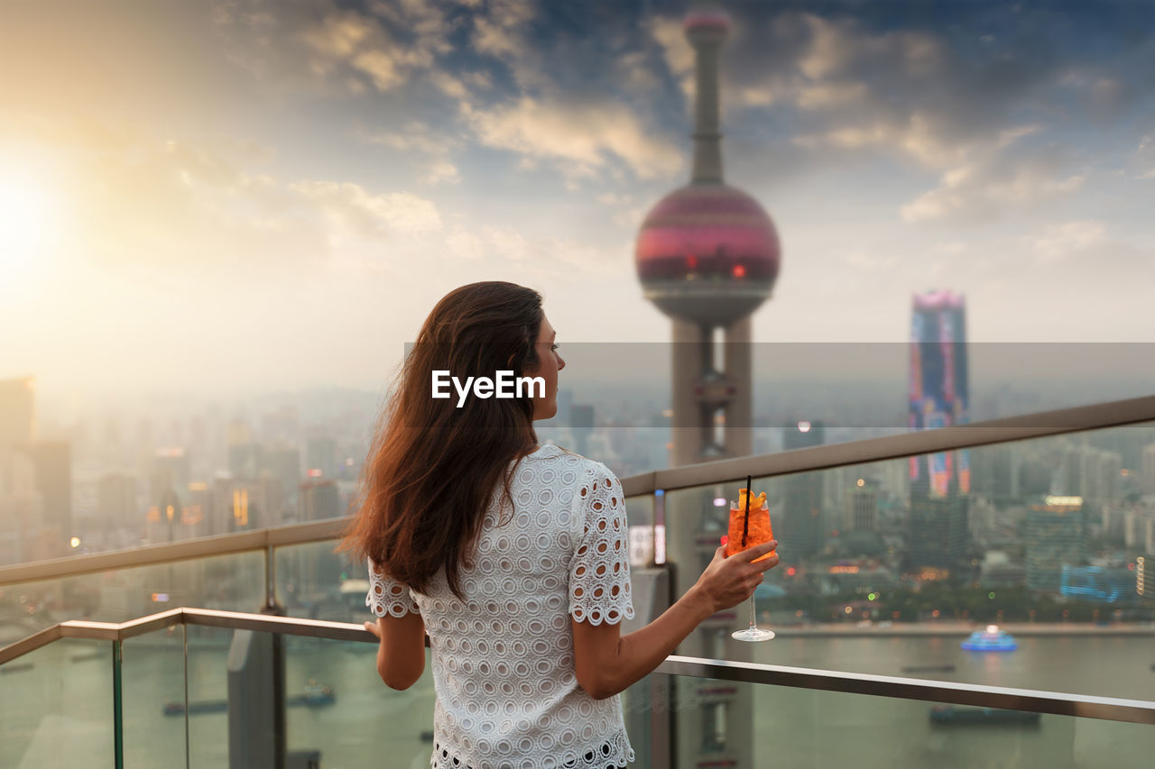 Rear view of mid adult woman looking at cityscape while standing by railing against sky during sunset