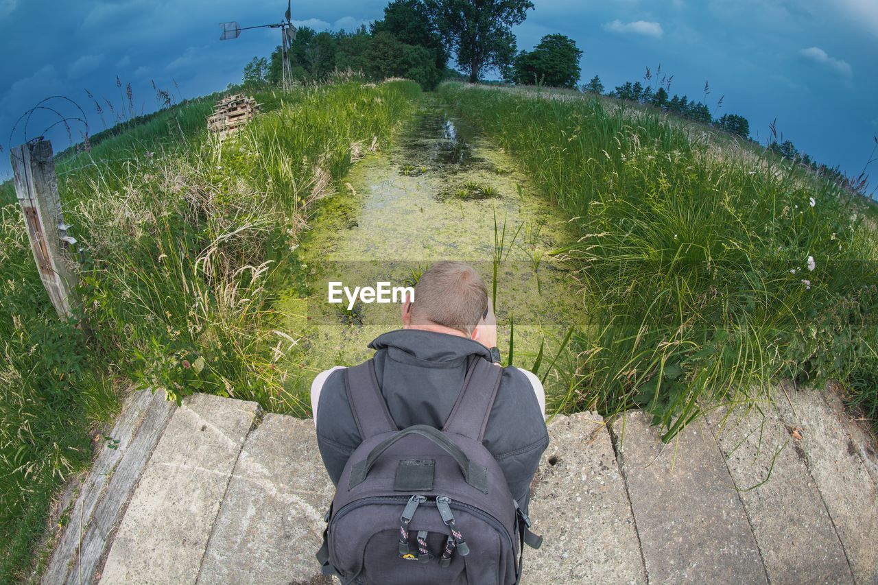 Rear view of man sitting on footbridge over stream at field