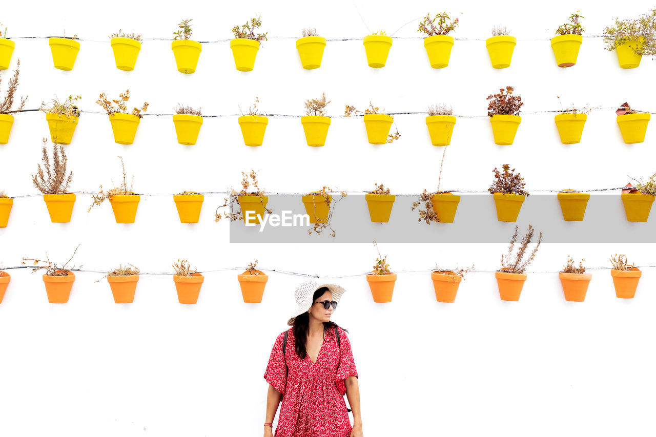 Cheerful female tourist in straw hat standing near wall with assorted plants in pots in rhodes on white background