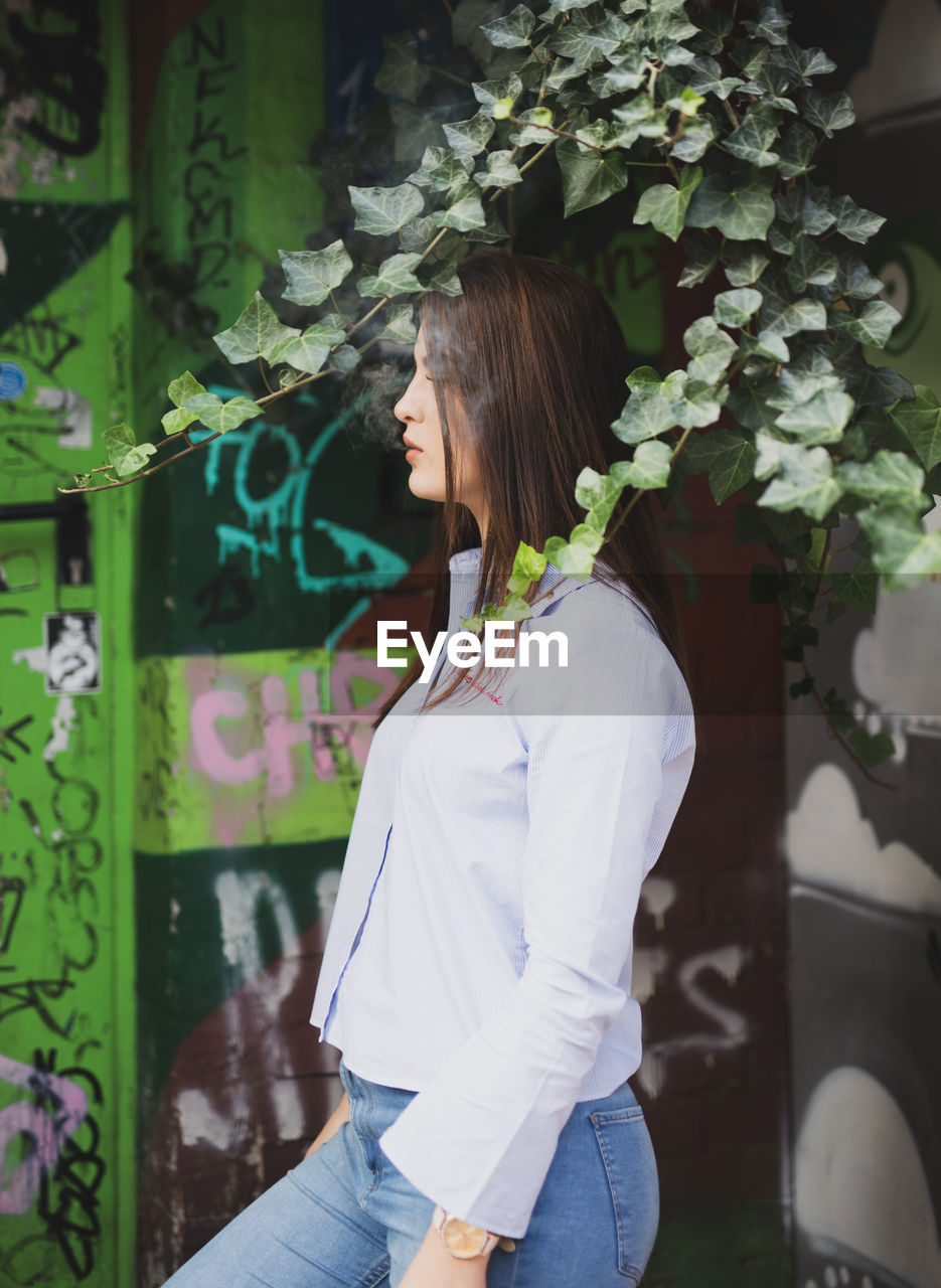 Profile view of young woman smoking while standing against graffiti wall