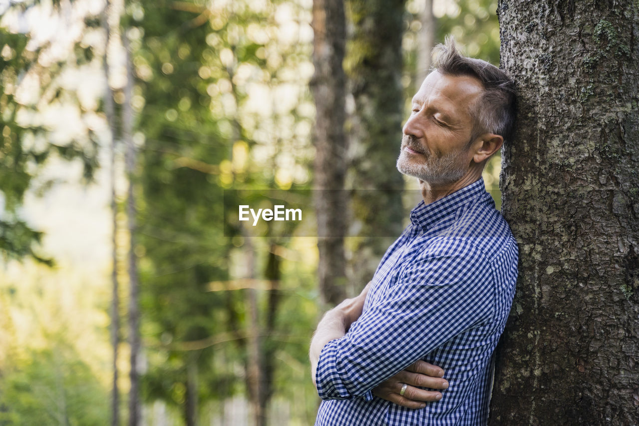 Mature man with eyes closed leaning on tree at forest