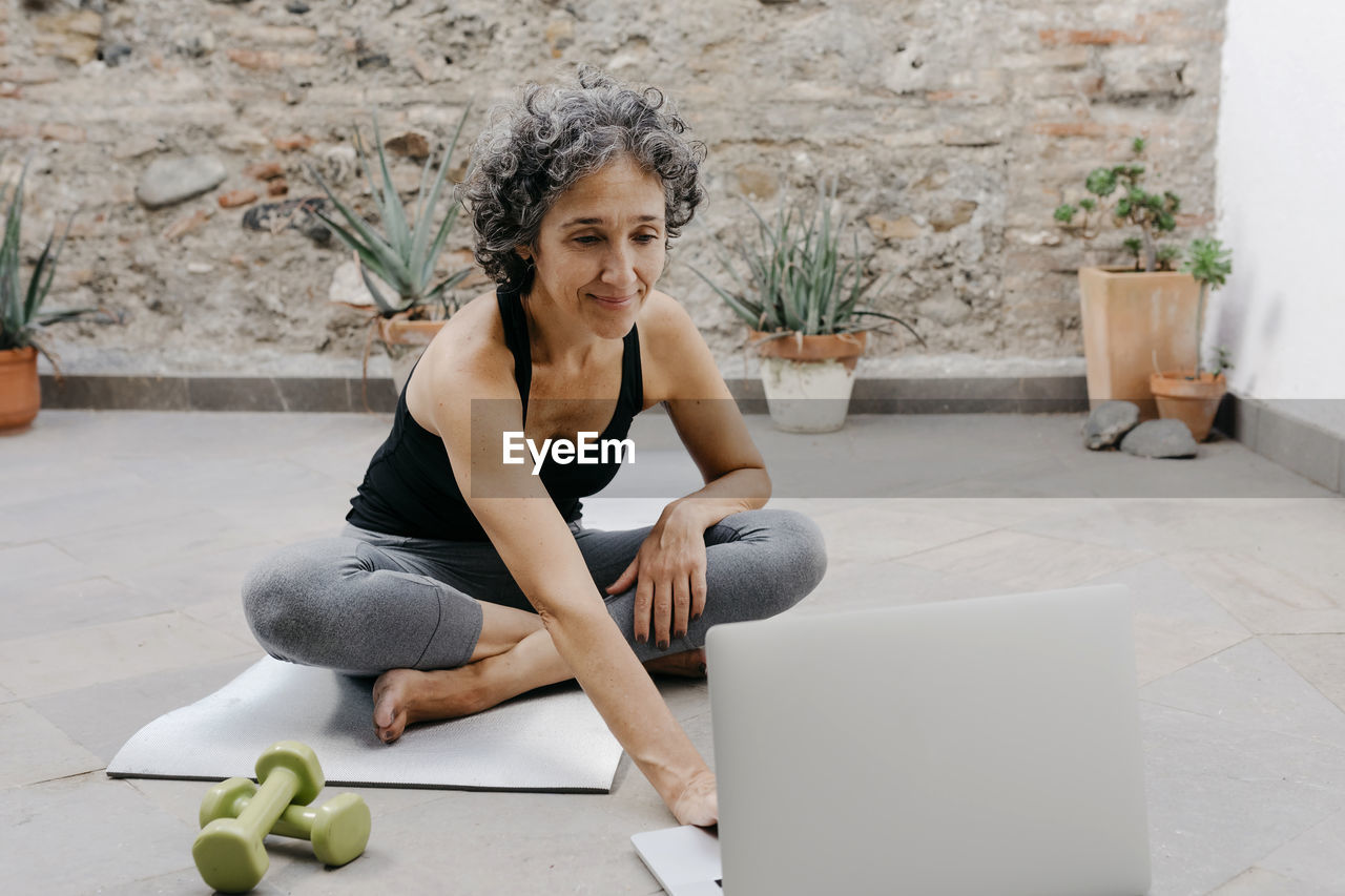 Smiling woman using laptop for learning yoga through online tutorial while sitting at back yard