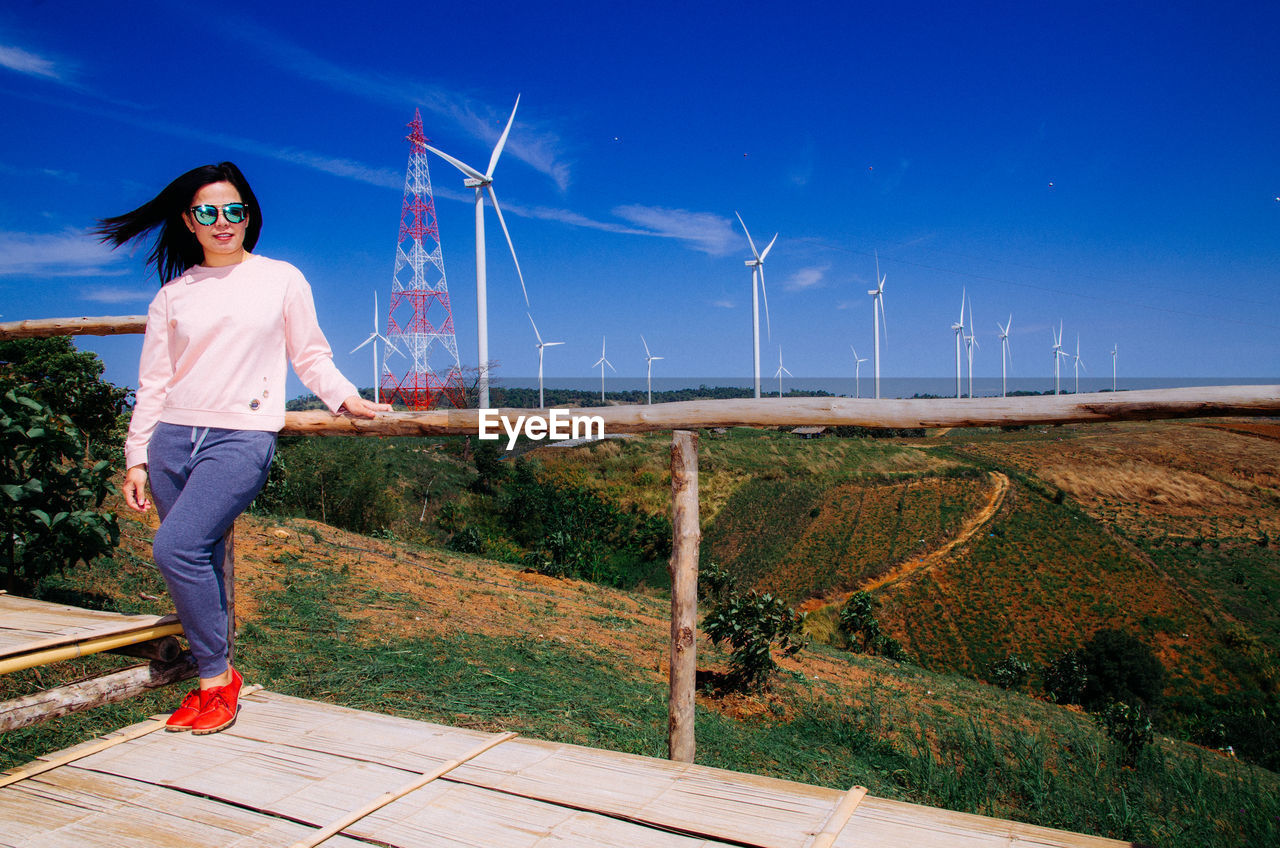 Full length portrait of young woman standing against wind turbines
