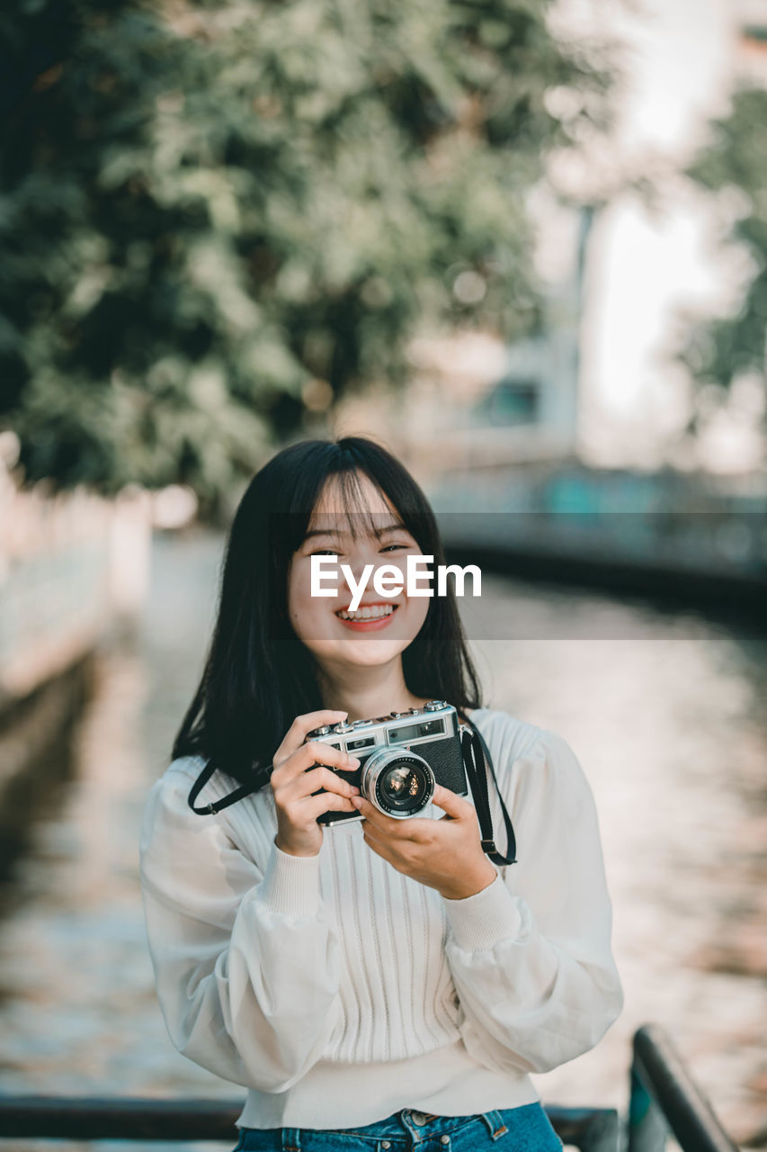 Portrait of smiling young woman holding camera standing by canal