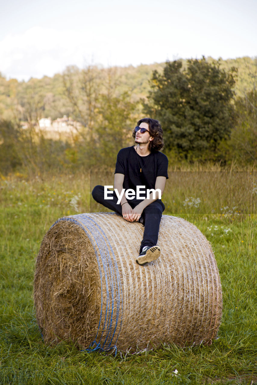 Full length of young man wearing sunglasses while sitting on hay bale against clear sky