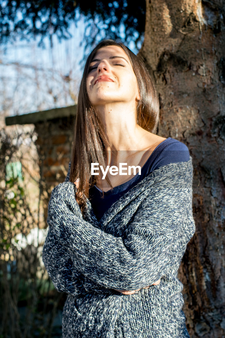 Young woman with closed eyes standing against tree during sunny day