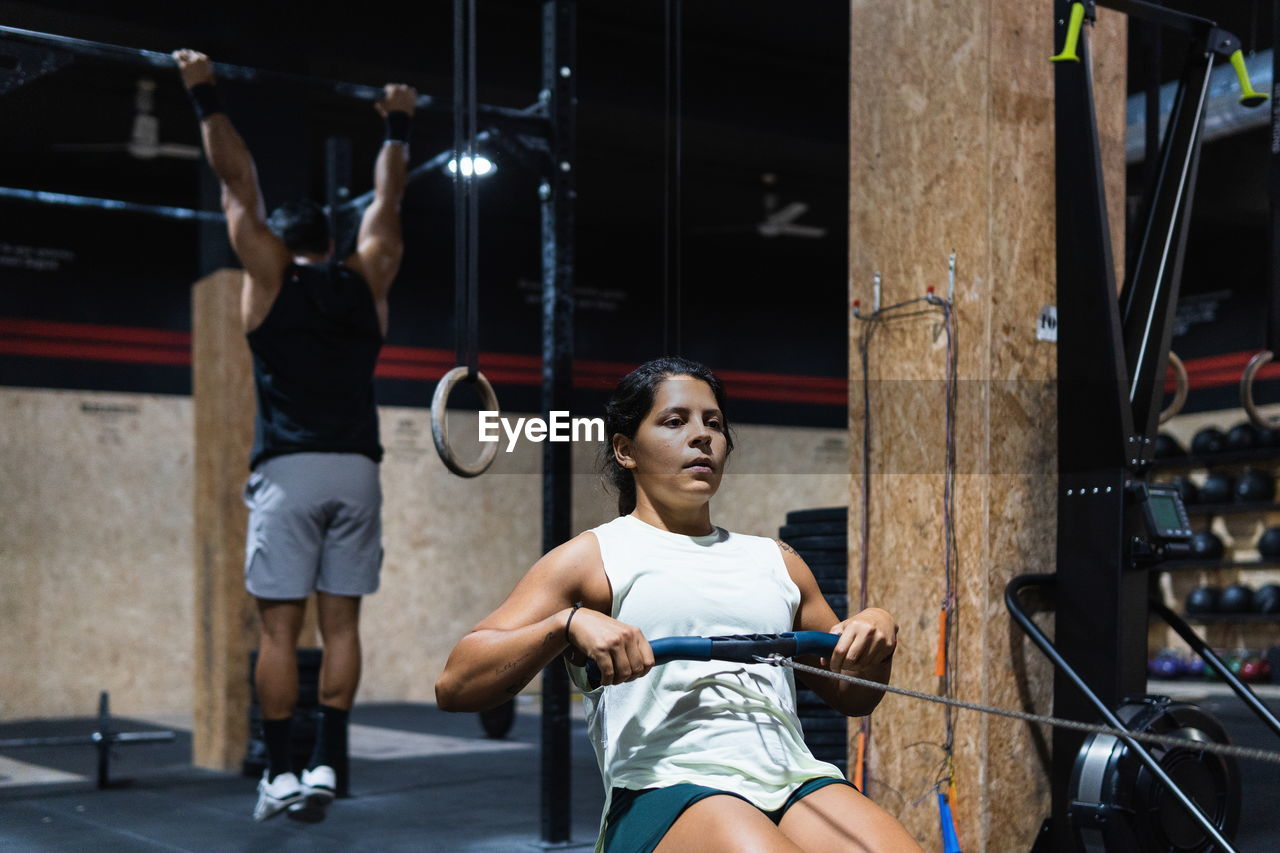 Strong hispanic sportswoman in activewear training on row machine during workout in light gym near sportsman handing on crossbar