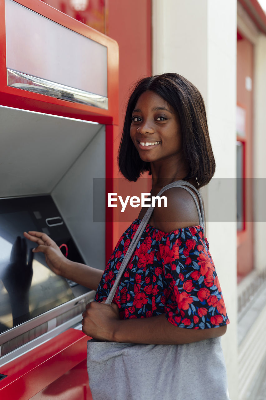 Smiling woman with shoulder bag standing at atm