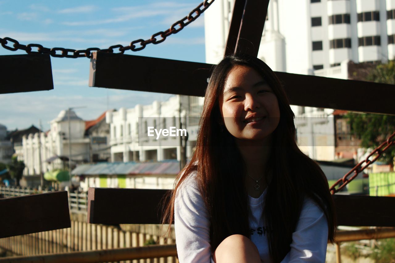 Portrait of smiling young woman sitting in city against sky