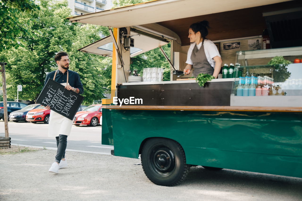 Male owner with blackboard looking at partner while walking by food truck