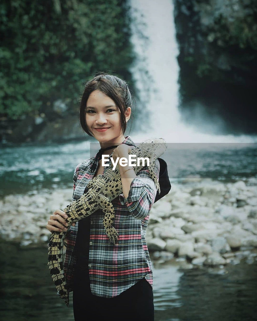 Portrait of smiling young woman holding crocodile against waterfall