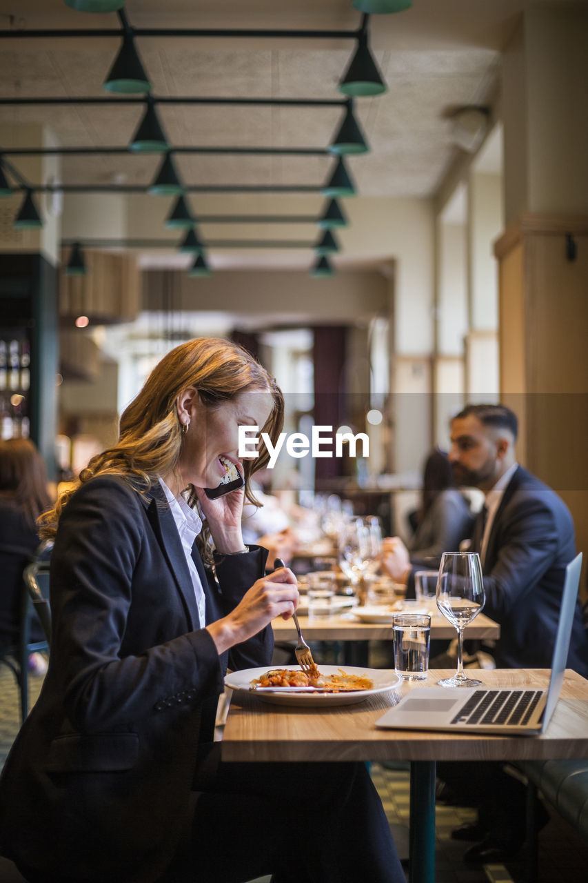 Smiling businesswoman talking on phone while eating in restaurant