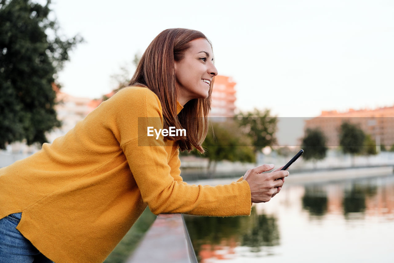 Smiling caucasian woman wearing yellow pullover using mobile phone device outdoors in park 