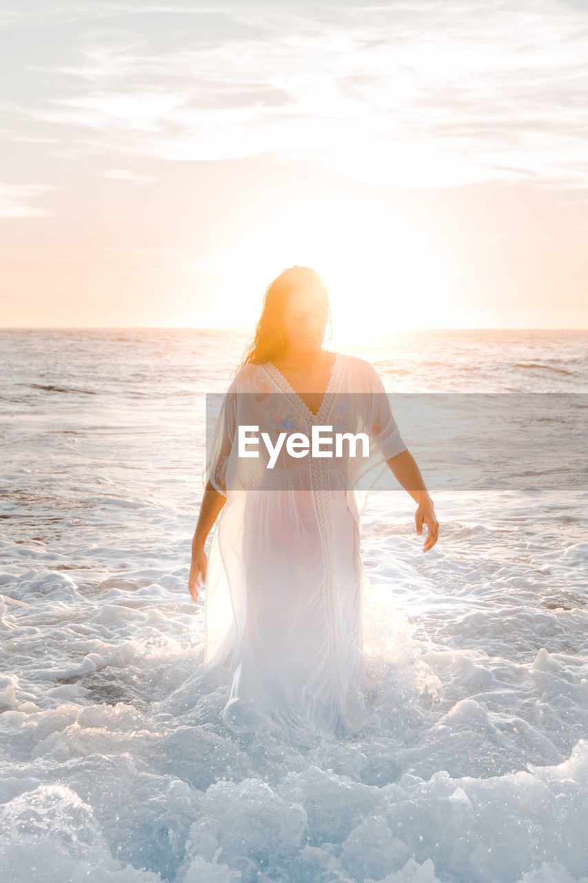 WOMAN STANDING AT BEACH DURING SUNSET