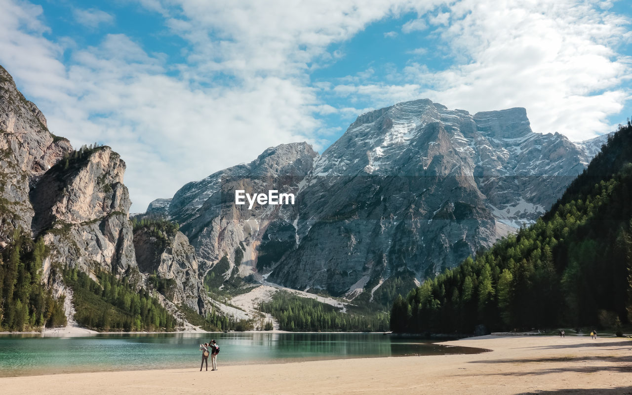 Rear view of couple standing by lake and mountains against cloudy sky