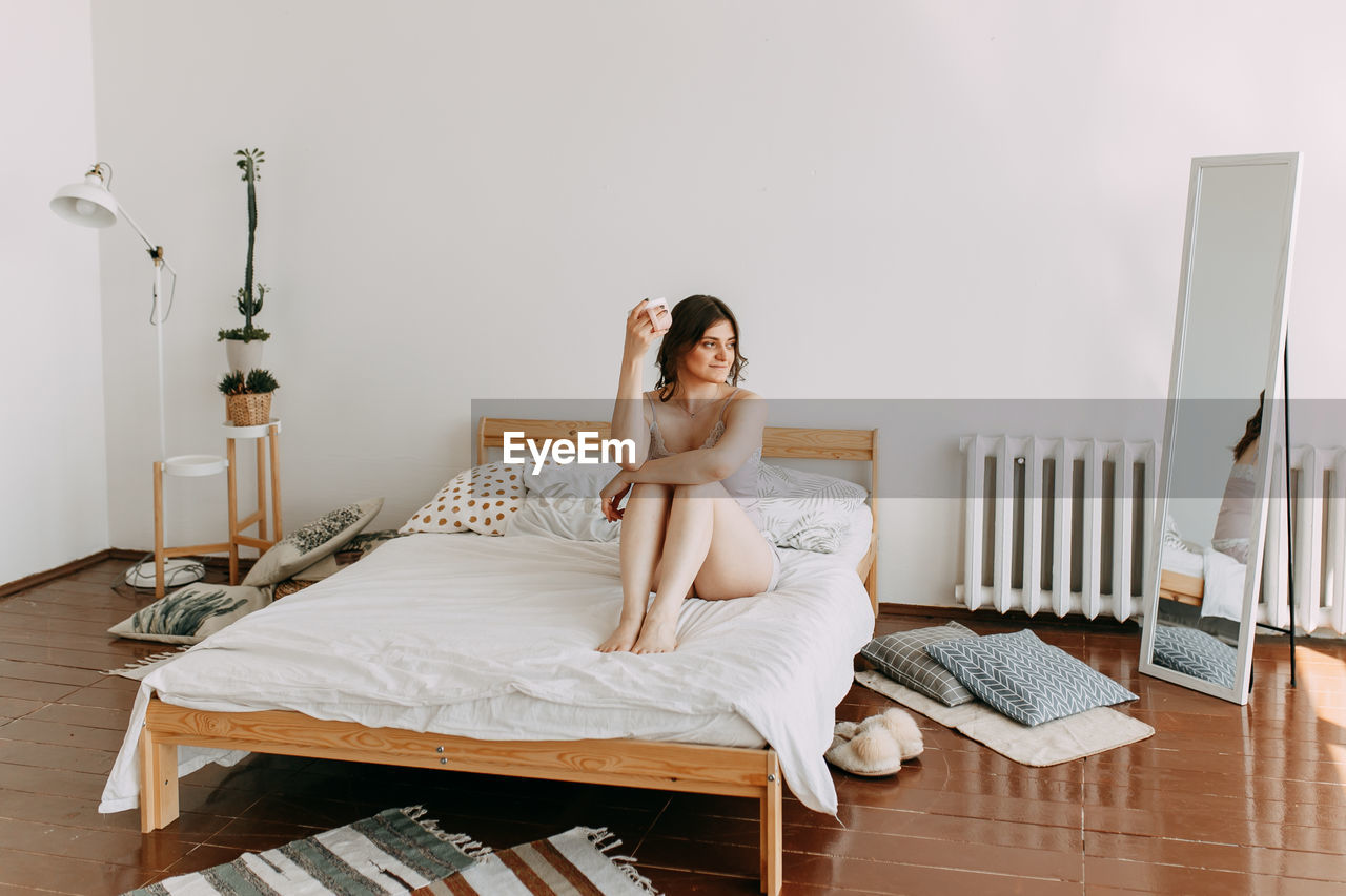 A happy girl in pajamas drinks coffee looks at the phone using technology while lying in bed at home