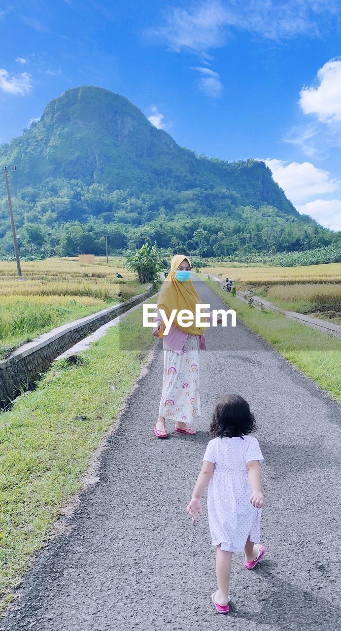 Rear view of mother wearing mask with daughter on road against sky