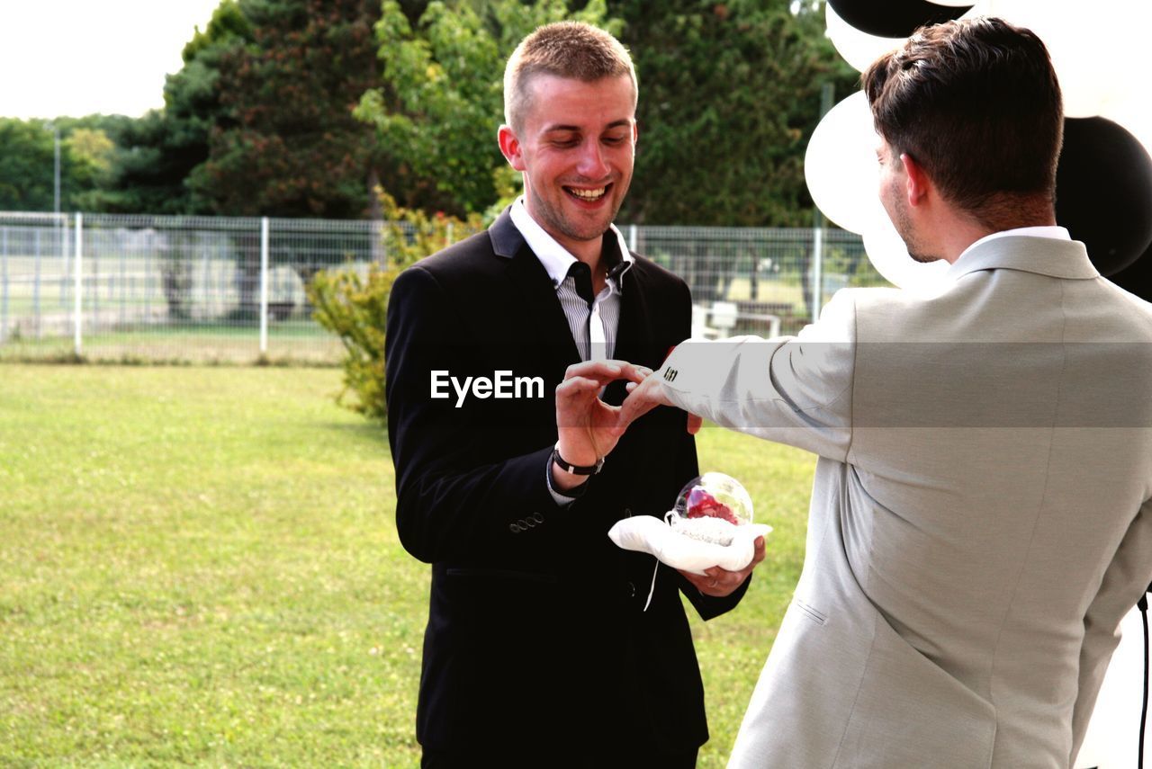 Cheerful man putting ring on boyfriend at park during wedding ceremony