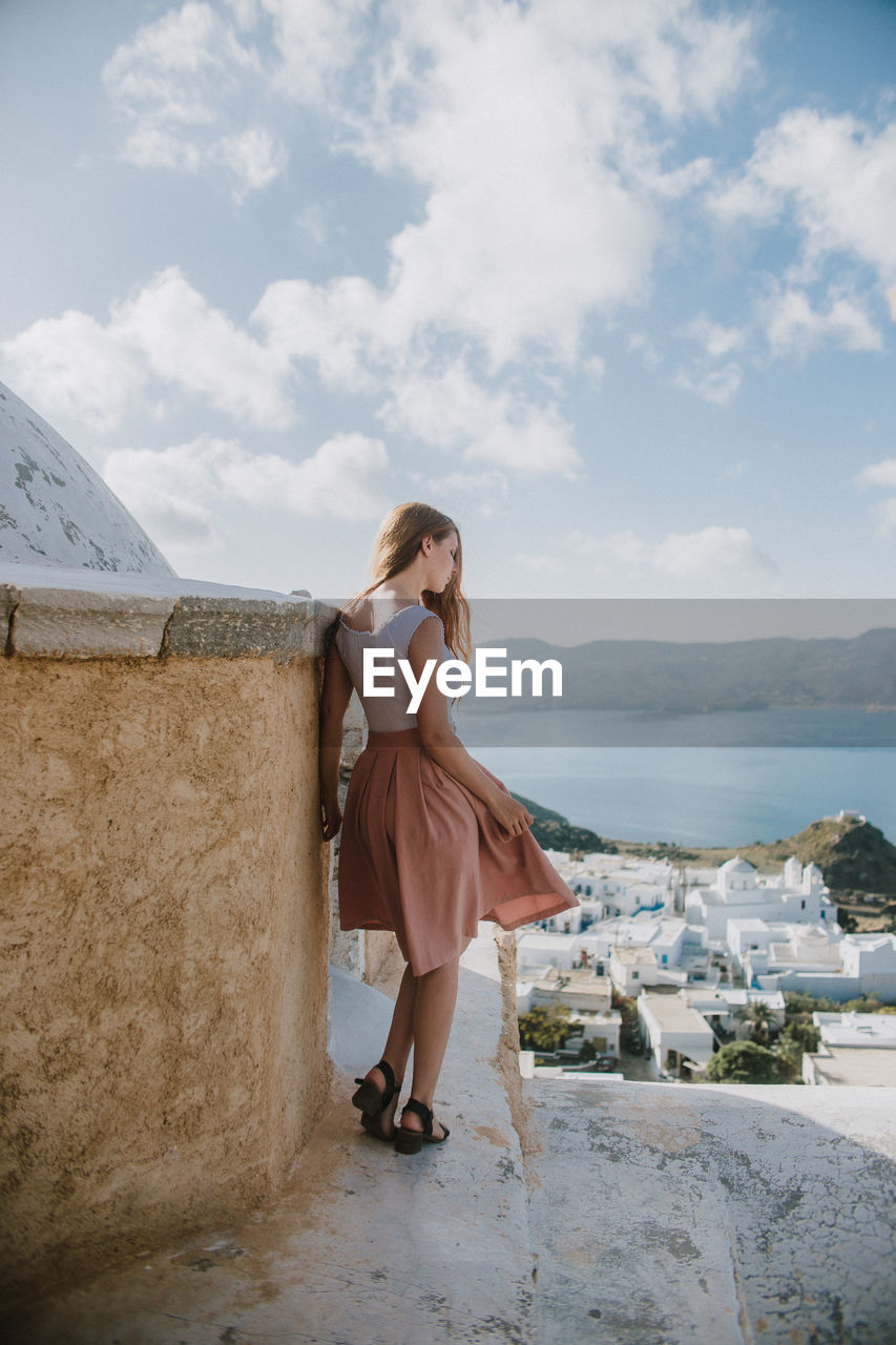 Back view anonymous slim female traveler in trendy sundress standing on shabby stone stairs in small coastal village with white cottages on sunny weather in greece