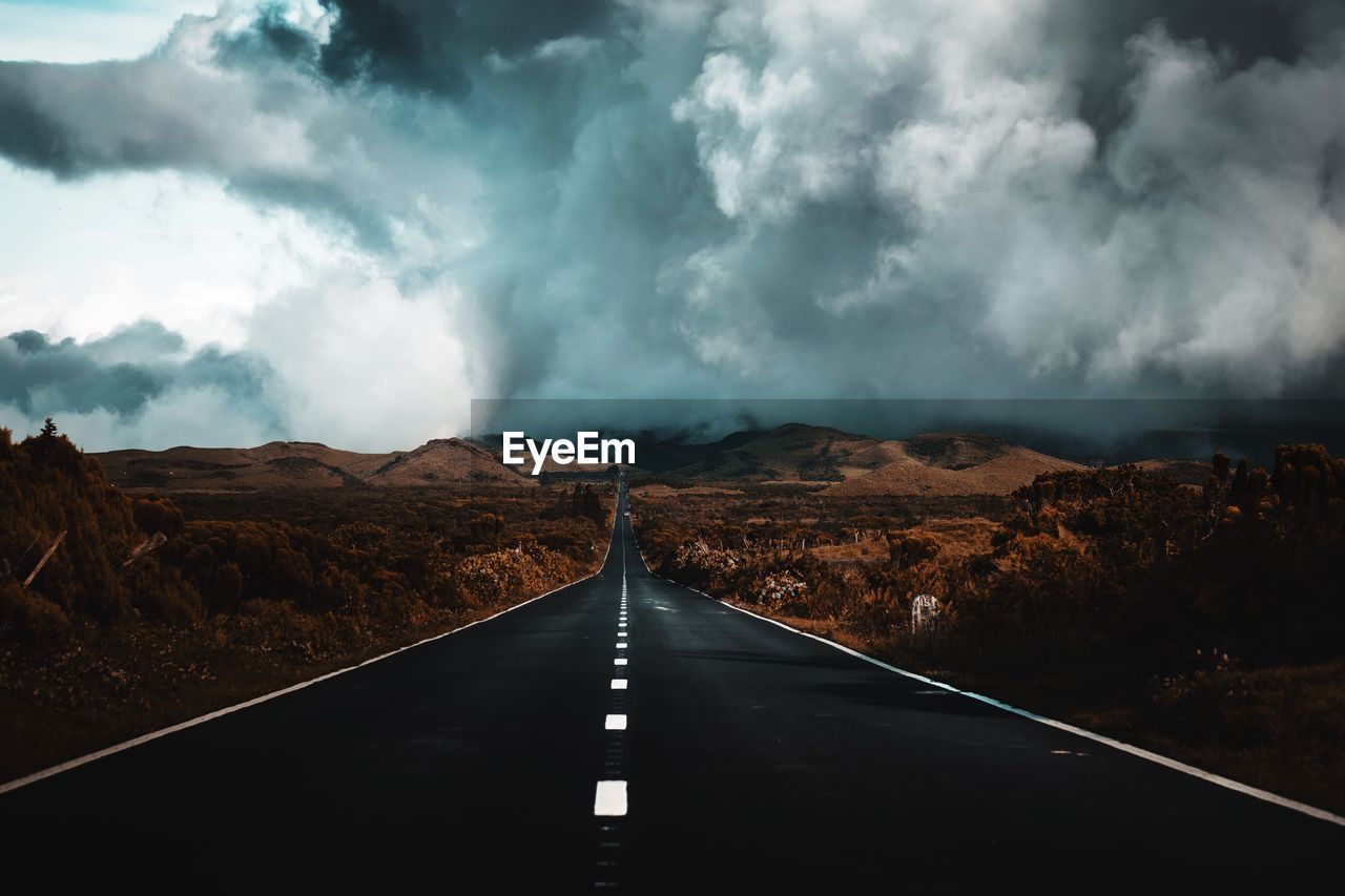 Diminishing perspective of empty road passing landscape against cloudy sky