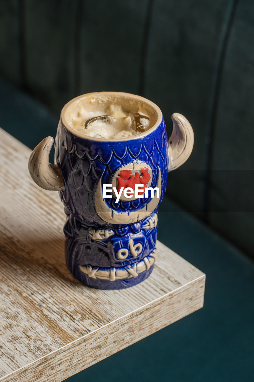 From above of bull shaped tiki mug of alcohol drink with froth placed against wooden table on blurred background