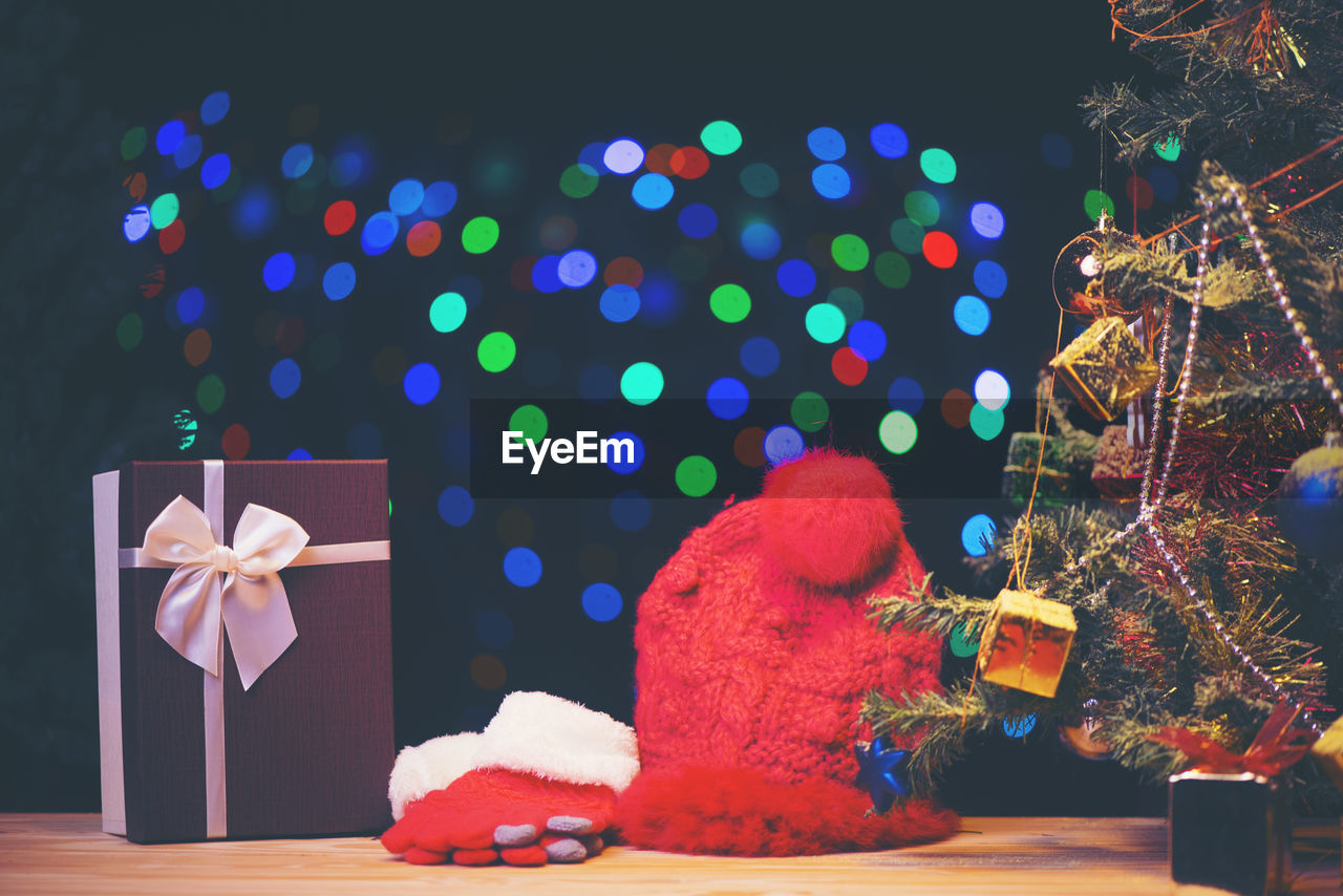 Close-up of knit hat and gloves by christmas tree against colorful lens flare