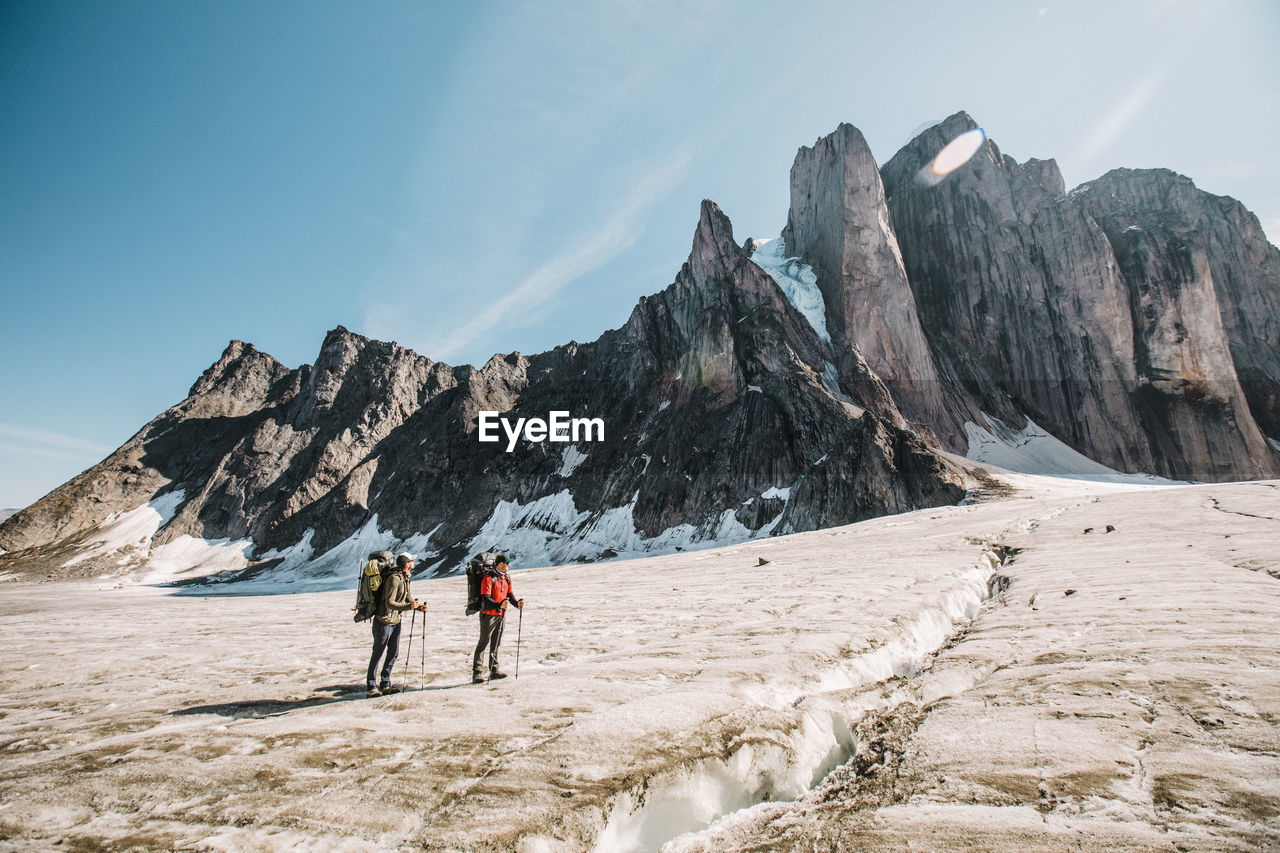 Two explorers standing on glcier below steep mountain summits.