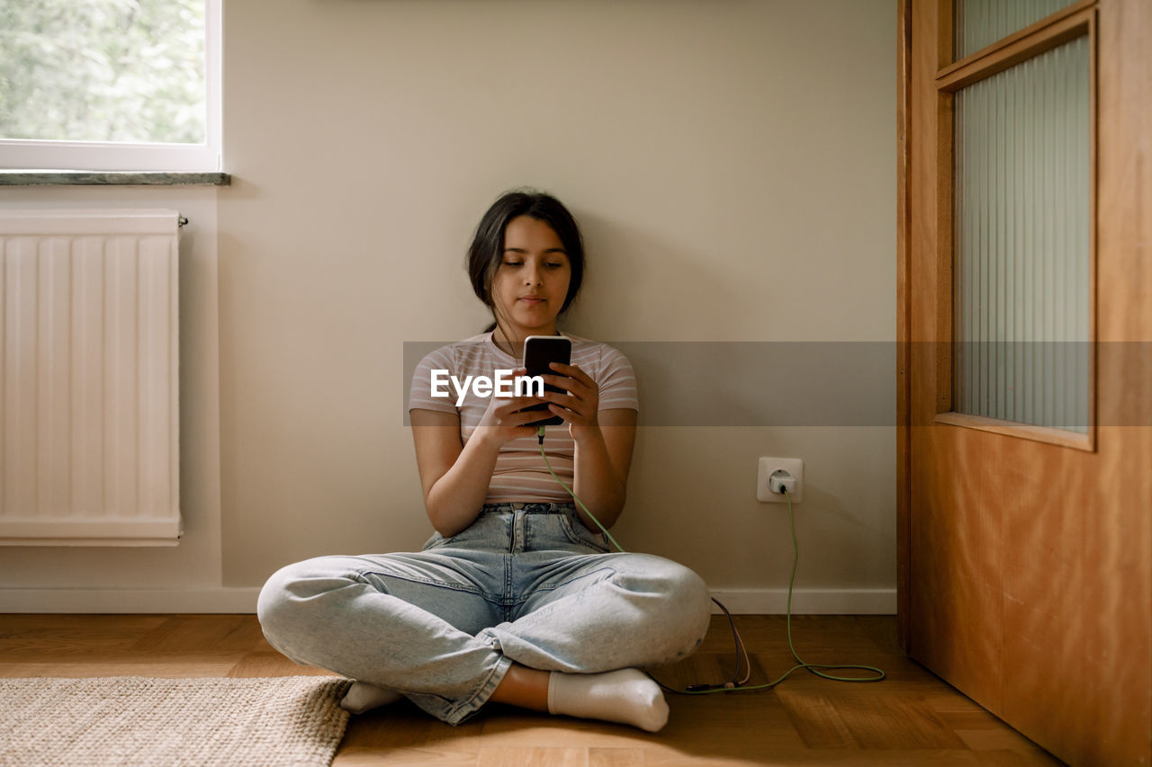 Girl using smart phone while sitting cross-legged on floor against wall at home