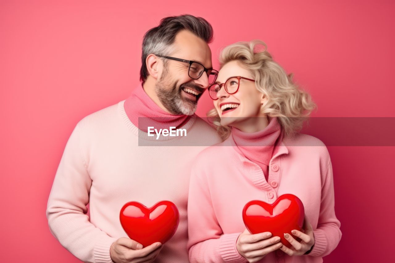 love, positive emotion, emotion, valentine's day, heart shape, heart, two people, romance, togetherness, adult, colored background, studio shot, red, happiness, men, women, kissing, smiling, affectionate, eyeglasses, indoors, glasses, portrait, pink, person, bonding, female, embracing, cheerful, young adult, passion, waist up, red background, fun, holding, human face, blond hair, joy, laughing, casual clothing, clothing, falling in love, facial hair