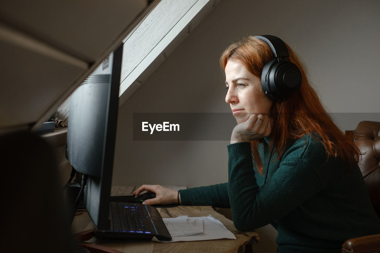 Middle age woman listening to music in big headphones near computer at home. creating play list