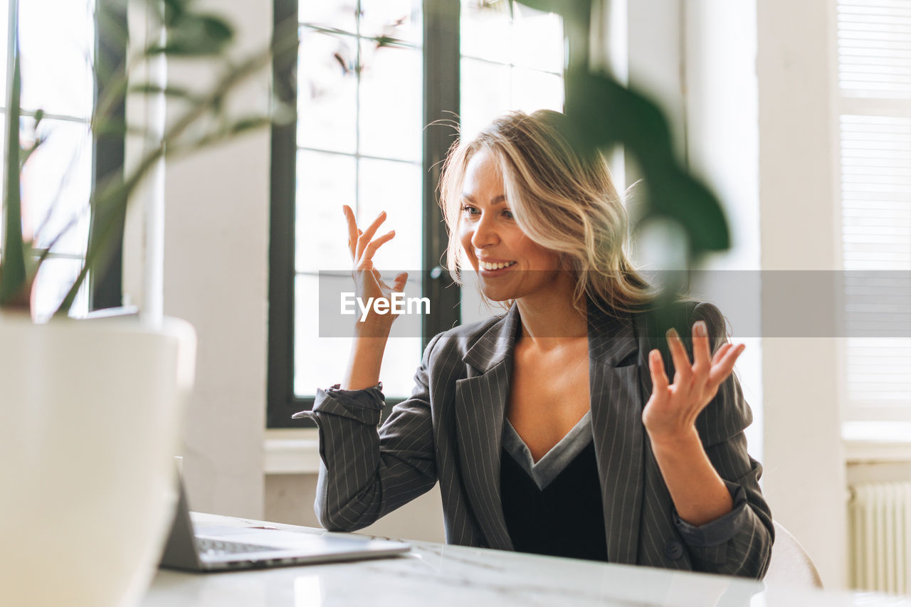 Young smiling blonde woman with long hair in stylish suit working at laptop in bright modern office