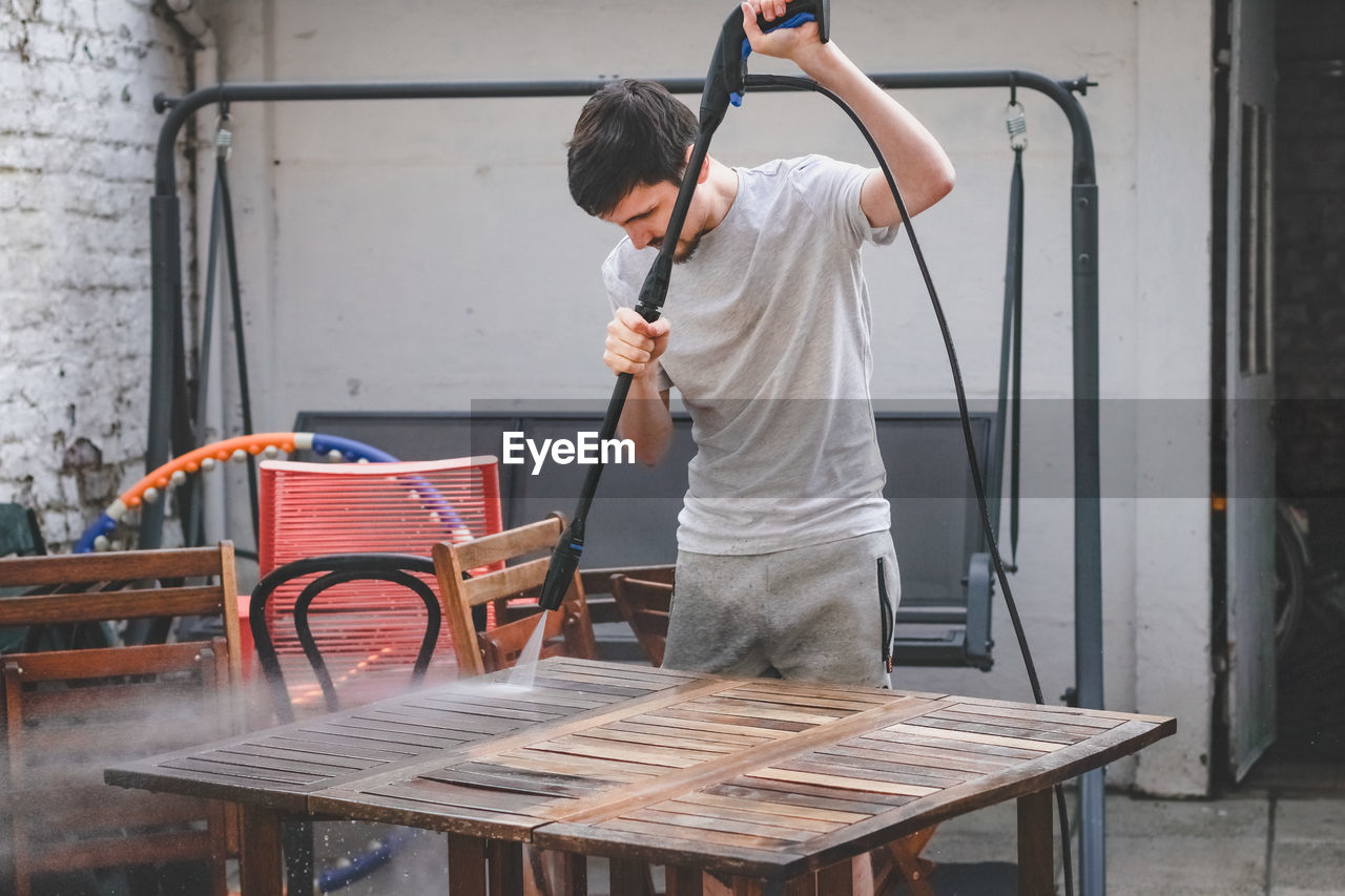 A caucasian guy in gray sweatpants and a t-shirt washes a wooden garden table with a karcher