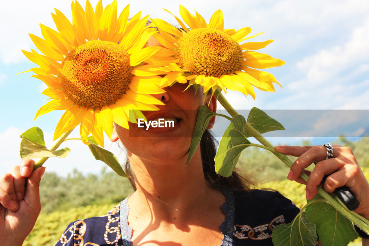 Caucasian brunette girl hides her eyes and part of her face behind two blooming yellow sunflower