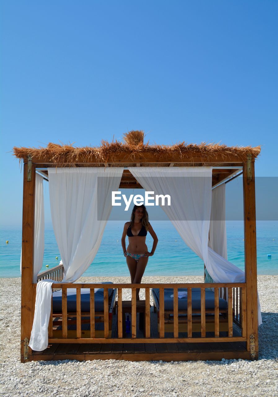 Woman wearing bikini standing at beach against clear blue sky