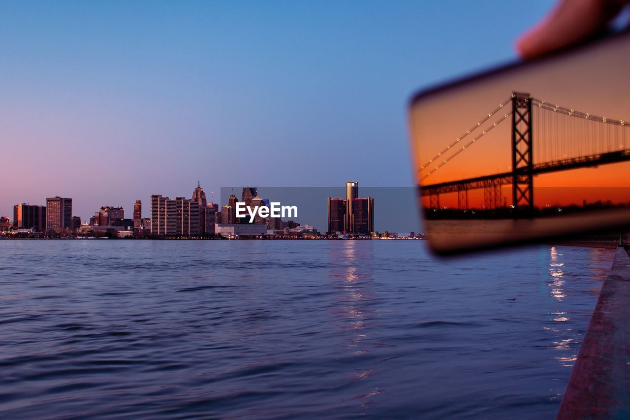 Bridge reflecting on mobile phone over river against clear blue sky during sunset