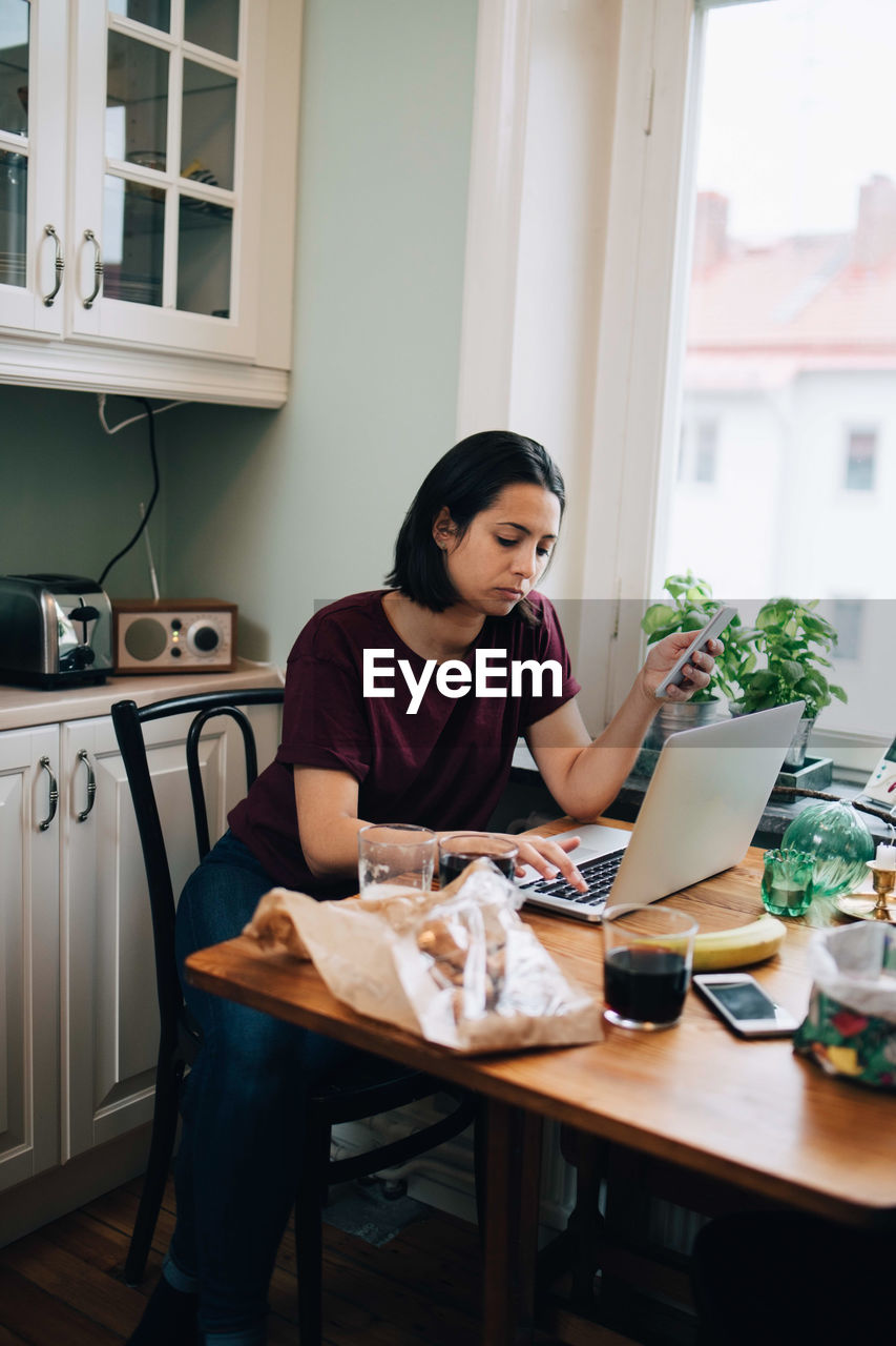 Woman using laptop while holding mobile phone at dining table in kitchen