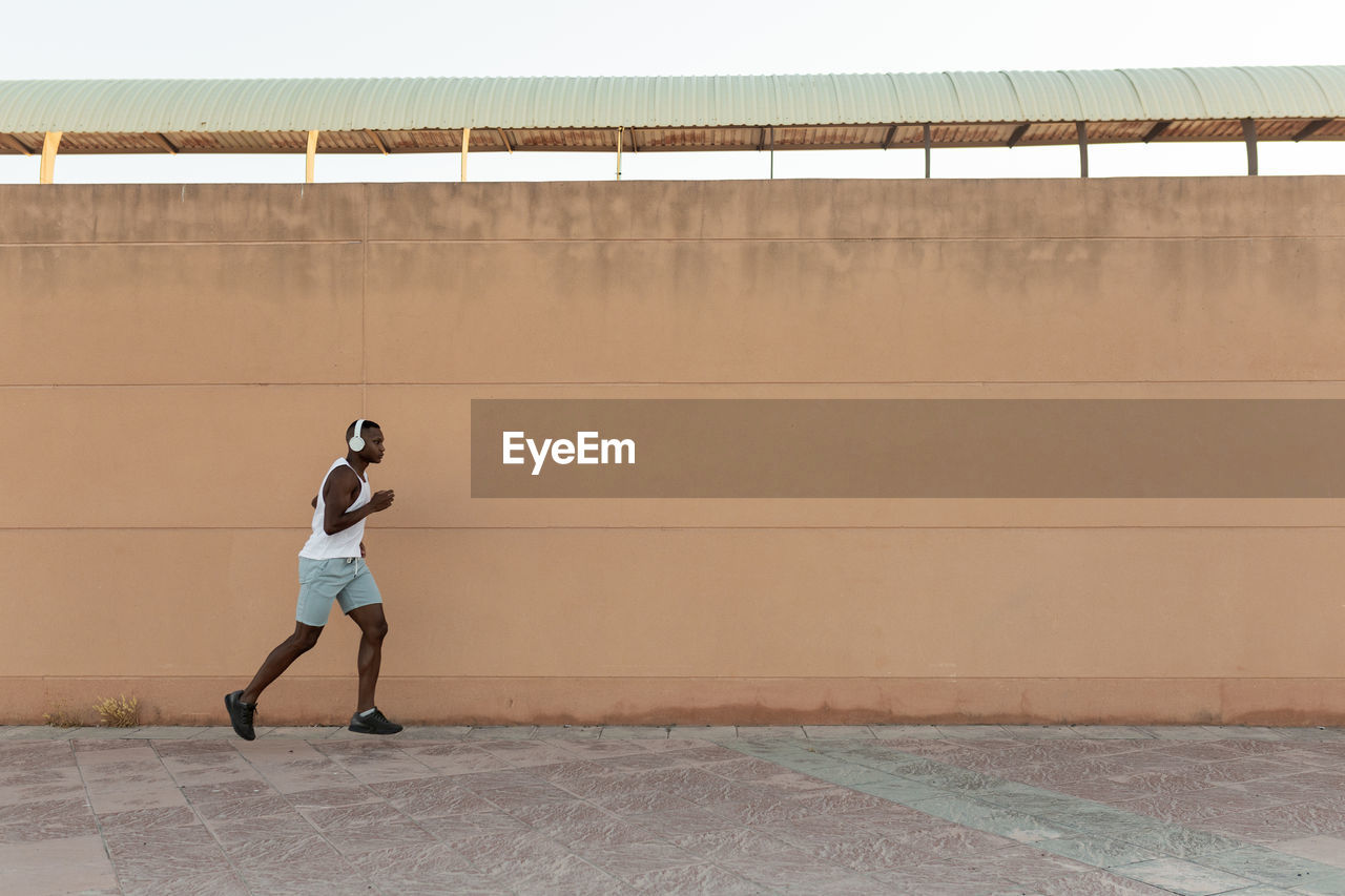 Side view full body of energetic african american sportsman in headphones jogging along pavement during fitness training