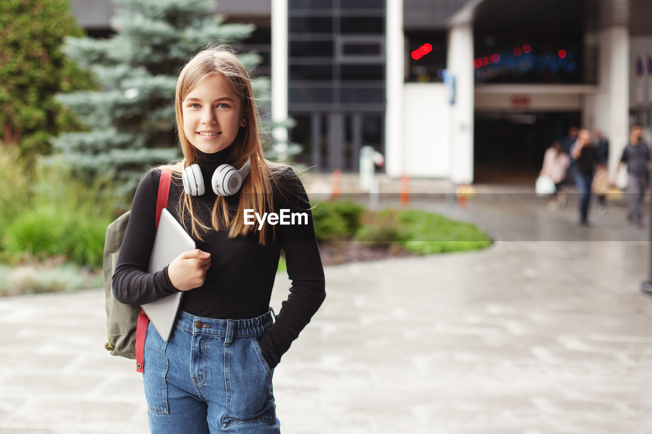 A beautiful fashionable female student in headphones stands in the city with a tablet and a backpack