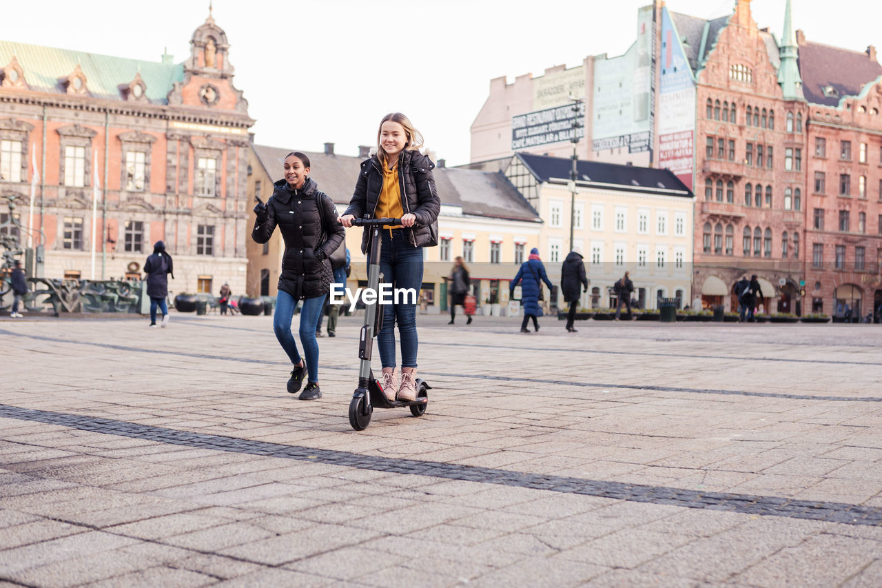 Friend walking behind teenage girl riding e-scooter on street in city