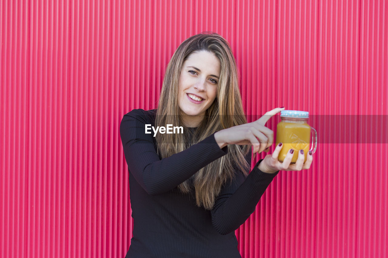 Portrait of smiling young woman holding drink in mason jar against wall