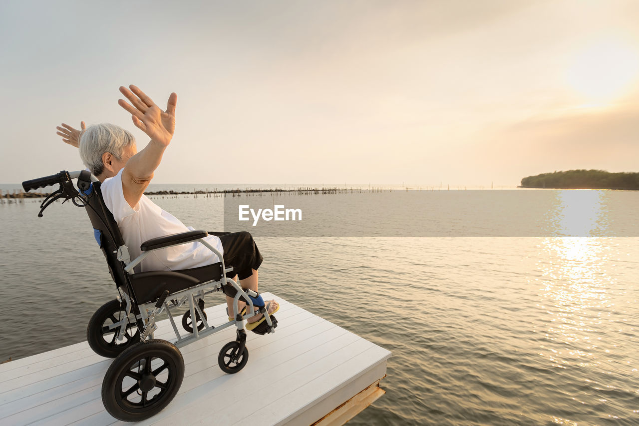 Side view of senior woman sitting on wheelchair against sea during sunset