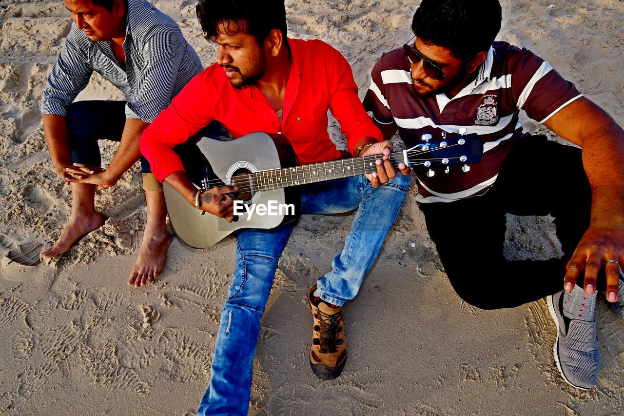 High angle view of man playing guitar while sitting with friends at sandy beach