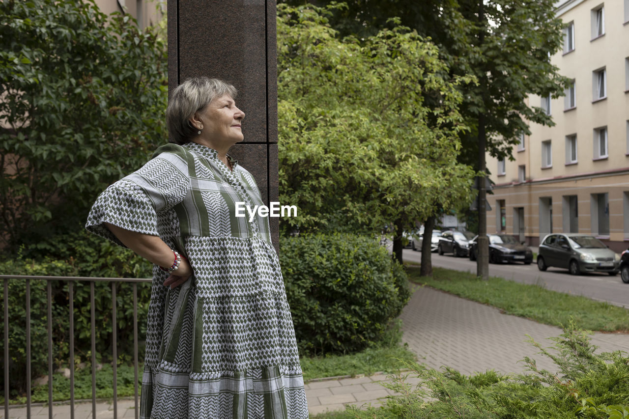 Real smiling female woman in yard of modern building. green trees around. copy space for text