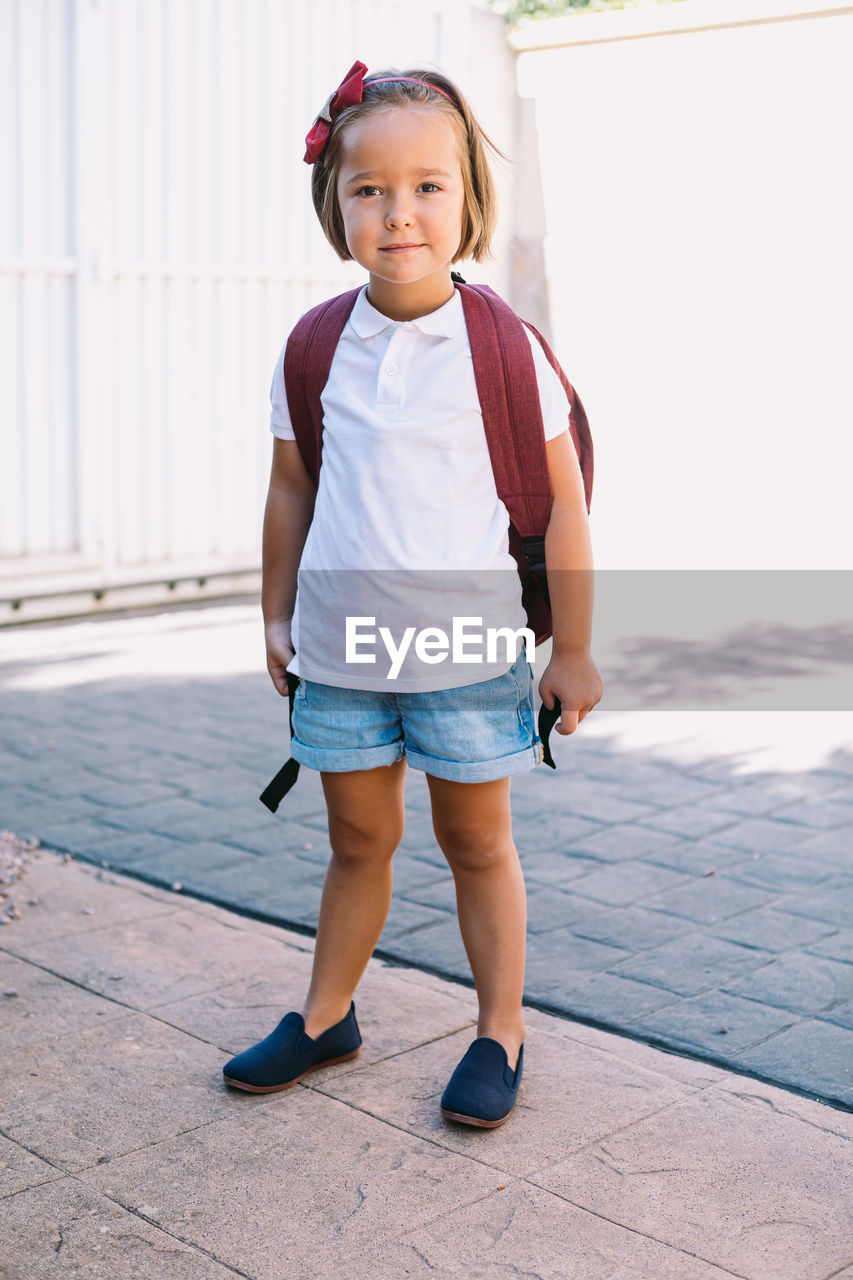 Schoolchild with backpack standing on pavement looking at camera in sunlight
