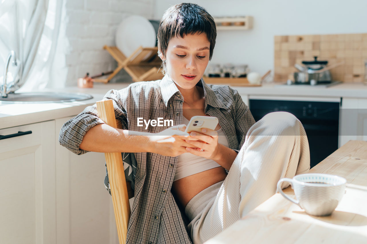 Young modern woman using phone for online messaging while sitting at home kitchen