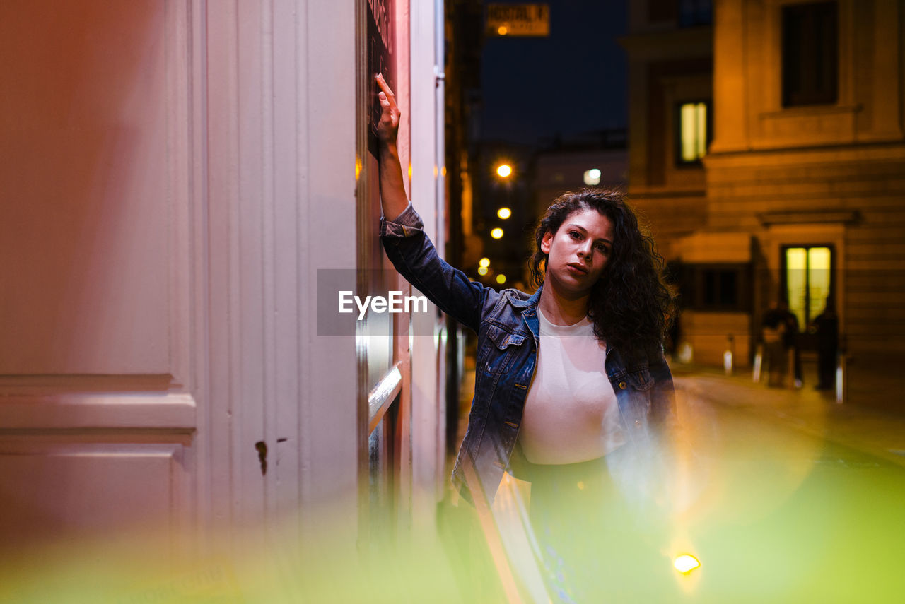 Confident young woman standing by wall on illuminated street at night