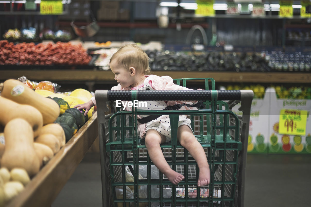 Full length of baby girl sitting on shopping cart while picking vegetables from market stall at supermarket